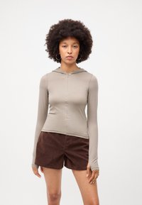 Langærmet, hættebluse i lys beige stof med lynlås foran. Sammen med brune, bløde shorts. Simpel, tætsiddende design.