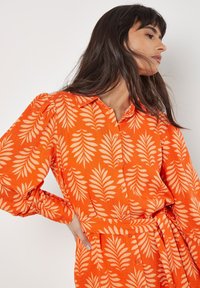 Oranges Kleid mit einem Muster aus weißen Blattformen, langen Ärmeln, einem kragenförmigen Ausschnitt und einem Taillenknoten.