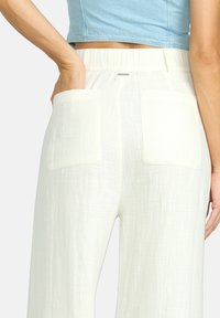 Vita linnebyxor med en strukturerad yta, som har stora bakfickor och ett elastiskt midjeband. En hand syns i en av fickorna.