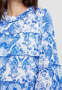 Blusa blanca con patrones florales y de paisley en azul. Presenta mangas en capas con volantes y un escote redondo. Tejido ligero.