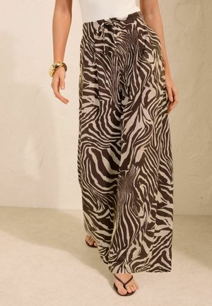 Kvinde iført brune og beige zebra-print vidde bukser med bindebælte, hvid top, guldbidé og brune sandaler, der går indendørs.
