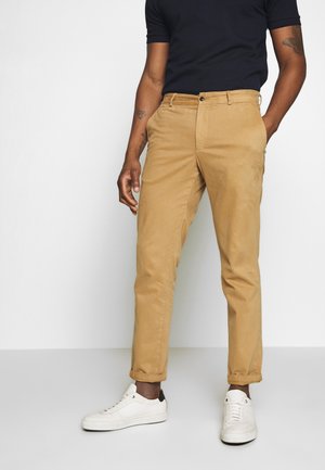 Mężczyzna noszący beżowe chinosy z podwiniętymi nogawkami, czarną koszulkę i białe trampki, stojący z jedną ręką w kieszeni na jednolitym białym tle.