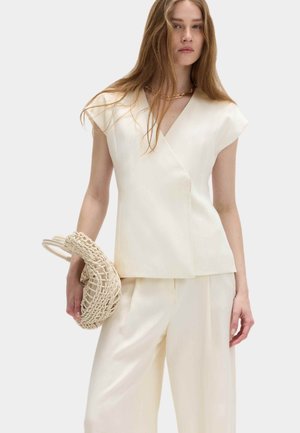 Kvinde med langt hår iført en off-white ærmeløs V-hals top og bukser med vide ben, der holder en flettet rund håndtaske i venstre hånd.