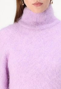 Lila pulover iz mehke, puhaste tkanine z visokim ovratnikom. Ima sproščen kroj in raglan rokave, kar dodaja k njegovi udobni teksturi.