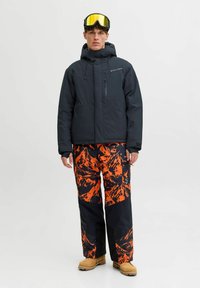 Juodas izoliacinis džempers, oranžinės ir juodos raštuotos sniego kelnės, geltonos akinių nuo saulės, ir smėlio spalvos batai, demonstruojami modelio prieš paprastą fono.