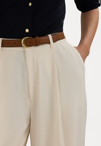 Beige geplooide broeken met een gladde textuur, voorzien van een bruin leren riem met een gouden gesp. Zwart shirt deels zichtbaar boven de broeken.