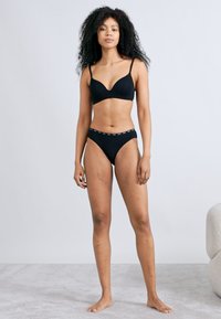Sort bikini-sæt lavet af glat stof. Bh'en har tynde stropper og en v-udskæring, mens trussen har et højt skåret design med et mønstret linning.