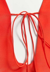 Gros plan d'un vêtement rouge avec des fines bretelles nouées en nœud sur un fond blanc.