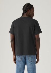 Μαύρο βαμβακερό T-shirt με άνετη εφαρμογή και κοντά μανίκια, με απλό σχέδιο και ίσια ρεβέρ. Πλάγια όψη, φορεμένο με μπλε τζιν.