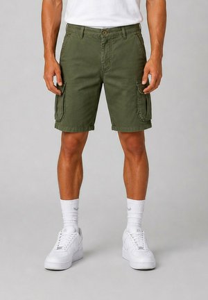Homme portant un short cargo vert olive, des chaussettes blanches et des baskets blanches, debout devant un fond gris uni.