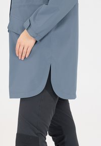 Ljusblå vattentålig kappa med böjd fåll, med en sidofrakturdetaill. Bärs över svarta åtsittande byxor. Slät textur, skräddarsydd passform.