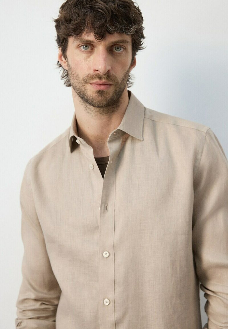 Homme aux cheveux bouclés bruns et aux yeux verts portant une chemise beige boutonnée, sur un fond clair uni.