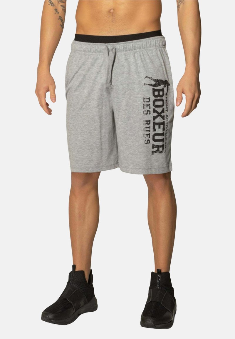 Graue Baumwollshorts mit einem schwarzen Bund, verziert mit fettem schwarzen Text an der Seite. Getragen mit schwarzen Sportschuhen. Einfaches Design, lockere Passform.