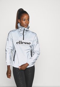Reflektierende silberne Jacke mit hohem Kragen und halbem Reißverschluss. Verfügt über ein großes schwarzes 'ellesse'-Logo und seitliche Reißverschlusstaschen.
