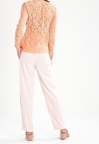 Perzikkleurige top met lange mouwen en kantoverlay, versierd met een ingewikkeld bloemenpatroon, gecombineerd met lichtroze wijde broekspijpen en beige block-heeled sandalen.