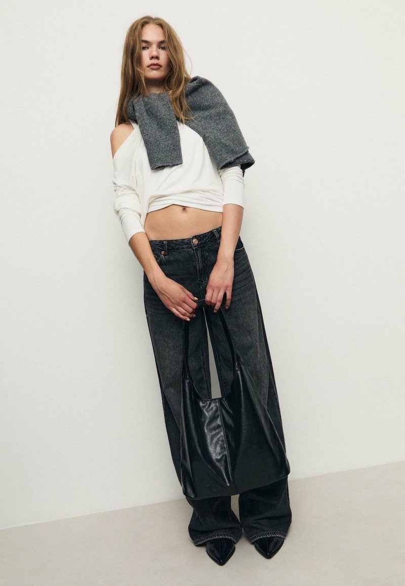 Top bianco off-shoulder, sciarpa grigia drappeggiata, jeans neri a gamba larga, in mano una borsa a tracolla nera in pelle, tacchi alti neri a punta.