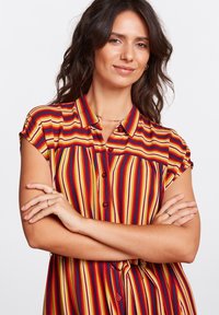 Camicia a righe con colletto, maniche corte arrotolate, bottoni sul davanti e una combinazione di colori rosso, giallo e blu.