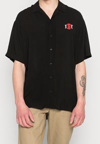 Camicia nera a maniche corte con bottoni e una vestibilità comoda, caratterizzata da un piccolo logo rosso e bianco di Tokyo sul lato sinistro del petto. Tessuto dalla superficie liscia.