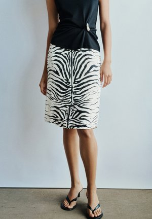 Femme portant un top noir torsadé, une jupe imprimée zèbre noire et blanche jusqu'aux genoux, et des sandales noires à talons, debout sur un sol en béton.
