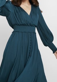 Teal omslagsklänning med långa puffärmar; smockad midja; och vertikala knapprader längs kjolen. Slät tygtextur.