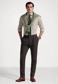 Herreantrekk med en rutete skjorte, grønn strikkegenser knyttet rundt skuldrene, mørkegrå dressbukser og brune skinnsko.