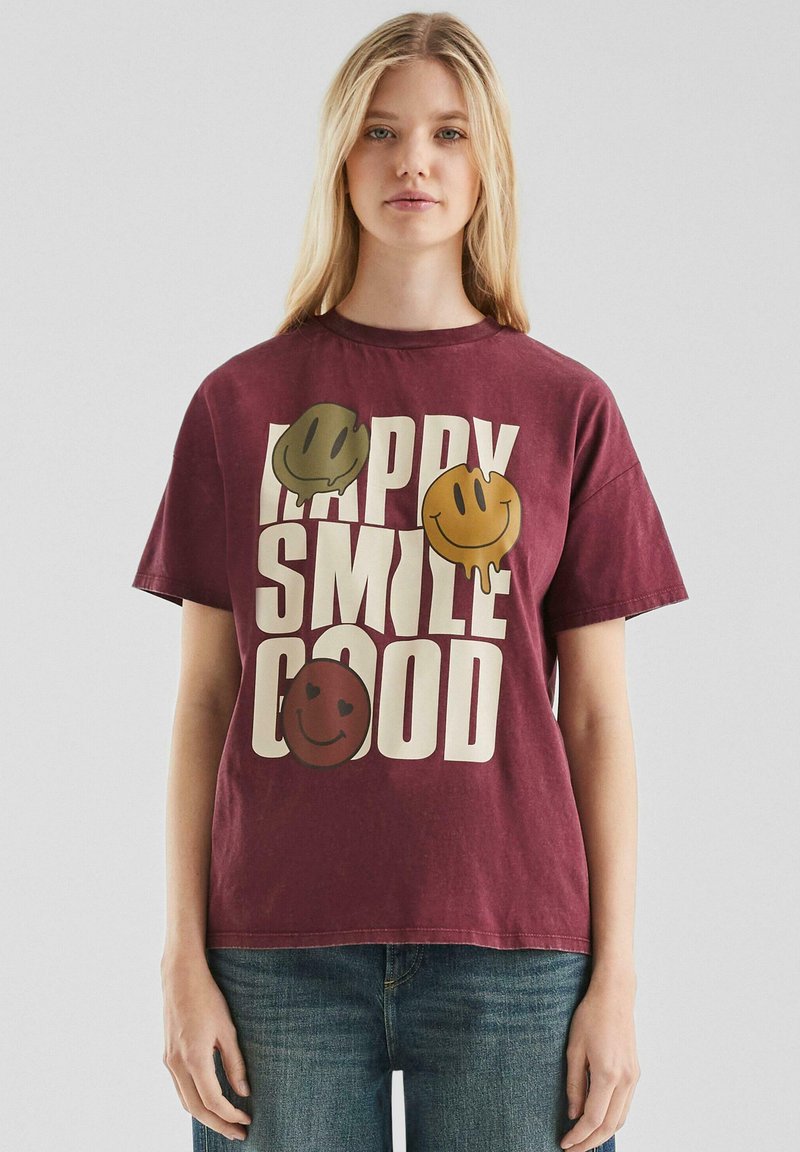 Jonge vrouw in een kastanjebruine T-shirt met beige tekst "HAPPY SMILE GOOD" en drie druipende smiley gezichten in groen, geel en bruin.
