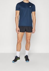 Marinefarbener kurzärmeliger Sportshirt mit einem Logo, kombiniert mit schwarzen Shorts, weißen Socken und schwarzen Laufschuhen mit grünen und blauen Akzenten.