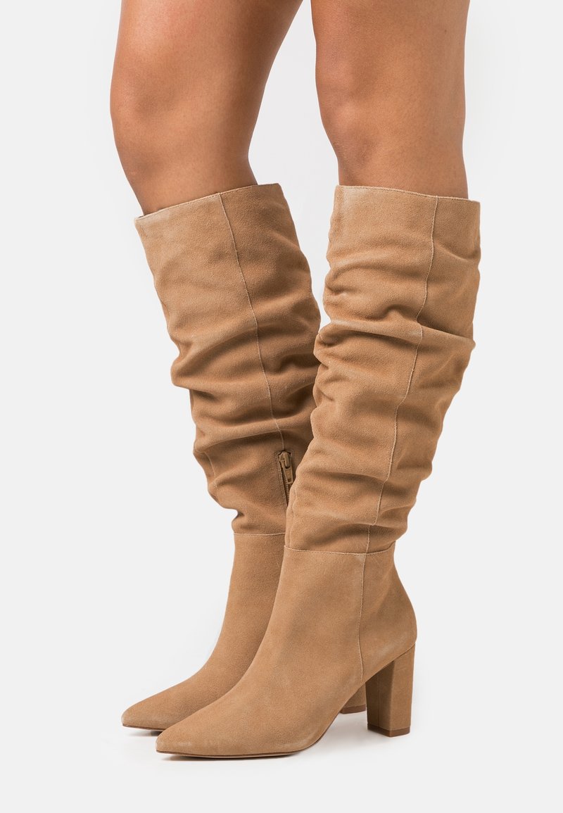 Bottes en daim beige au-dessus du genou avec un design décontracté, un bout pointu et un talon épais. Elles sont équipées d'une fermeture éclair latérale pour un enfilage facile.