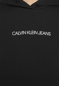 Sweat à capuche noir avec texte blanc « CALVIN KLEIN JEANS » centré sur la poitrine, montrant la texture du tissu et les détails des coutures.