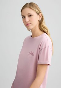 Tricou roz deschis cu mânecă scurtă, de culoare roz pal, cu guler rotund și un mic logo "LIUJO" într-o nuanță mai închisă. Material din bumbac moale.