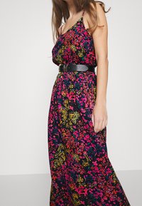 Blommig maxiklänning i mörk tyg med livliga rosa, gula och gröna mönster. Har tunna axelband och ett svart bälte i midjan.