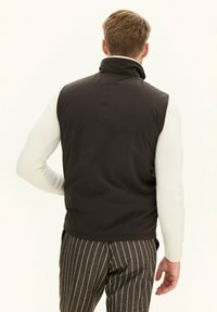Braune, ärmellose Weste mit hohem Kragen, aus glattem Material, kombiniert mit einem cremefarbenen Langarmhemd und gestreiften Hose.