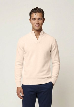 Młody mężczyzna uśmiechający się, ubrany w jasnobeżowy sweter z dekoltem w serek i granatowe spodnie, stojący na jednolitym tle.