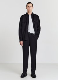 Schwarze Hemdbluse über einem hellgrauen T-Shirt, kombiniert mit maßgeschneiderter schwarzer Hose und schwarzen Lederschuhen. Schlichtes, elegantes Design.