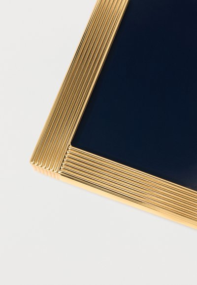 Λεπτομέρεια γωνίας ενός σκουρομπλε επιφάνειας πλαισιωμένης από ένα υφασμένο, χρυσό μεταλλικό πλαίσιο με παράλληλες ράβδους σε λευκό φόντο.