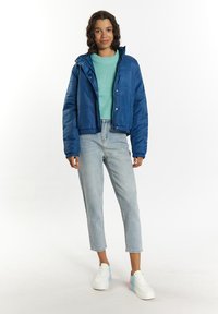 Blå vadderad jacka med huva, tillsammans med en ljusgrön tröja och ljusa denimjeans. Vita sneakers med gradientdetaljer.