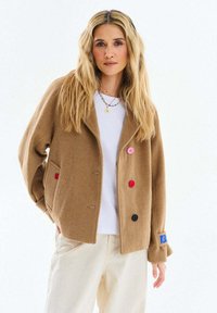 Blondína s dlhými vlasmi, oblečená v ťavej bunde s farebnými gombíkmi, bielej košeli, krémových nohaviciach a s náhrdelníkom z korálok na bielom pozadí.