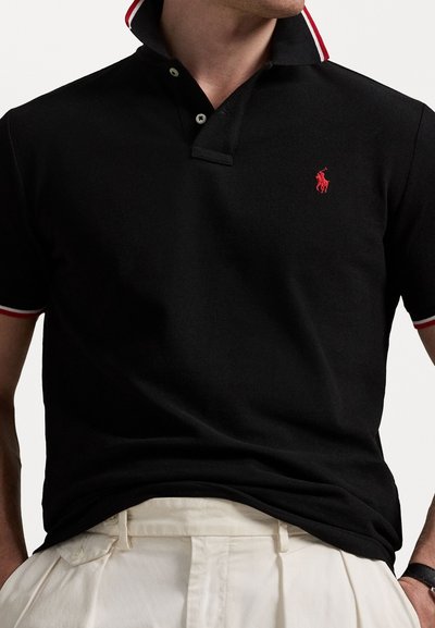 Homem a usar uma camisa polo preta com colarinho vermelho e branco e um logotipo de cavalo bordado em vermelho, acompanhado por calças brancas.