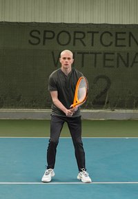 Egy férfi áll egy kék teniszpályán, narancssárga teniszütőt tart a kezében, sötét színű pólót és fekete nadrágot visel, fehér cipőben.