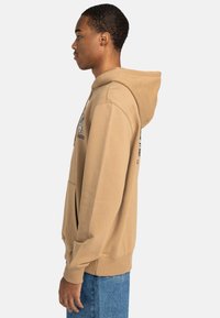 Brown hooded sweatshirt with a kangaroo pocket, ribbed cuffs, and a relaxed fit; features graphic design and printed text on the front.