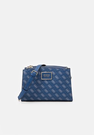 Blå Guess crossbody-taske med justerbar rem, mønstret med hvide logoer og et metalmærke foran med teksten "Guess Est 1981".