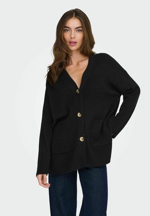Jeune femme aux longs cheveux bruns portant un cardigan noir oversize boutonné et un jean bleu foncé, posant avec une main dans la poche.