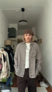 Outfit casual che presenta una camicia a quadri con bottoni, una maglietta bianca, pantaloni marroni e un berretto marrone; in piedi in una stanza con decorazioni minime.
