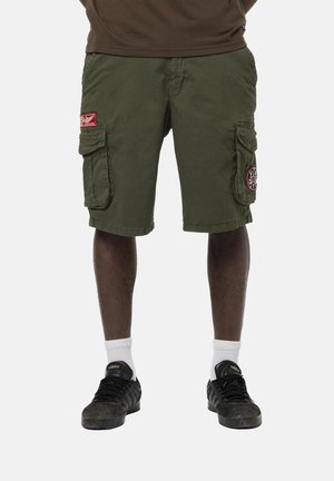 Homme portant un short cargo vert olive avec poches latérales, chaussettes blanches et baskets Adidas noires, debout contre un fond uni.