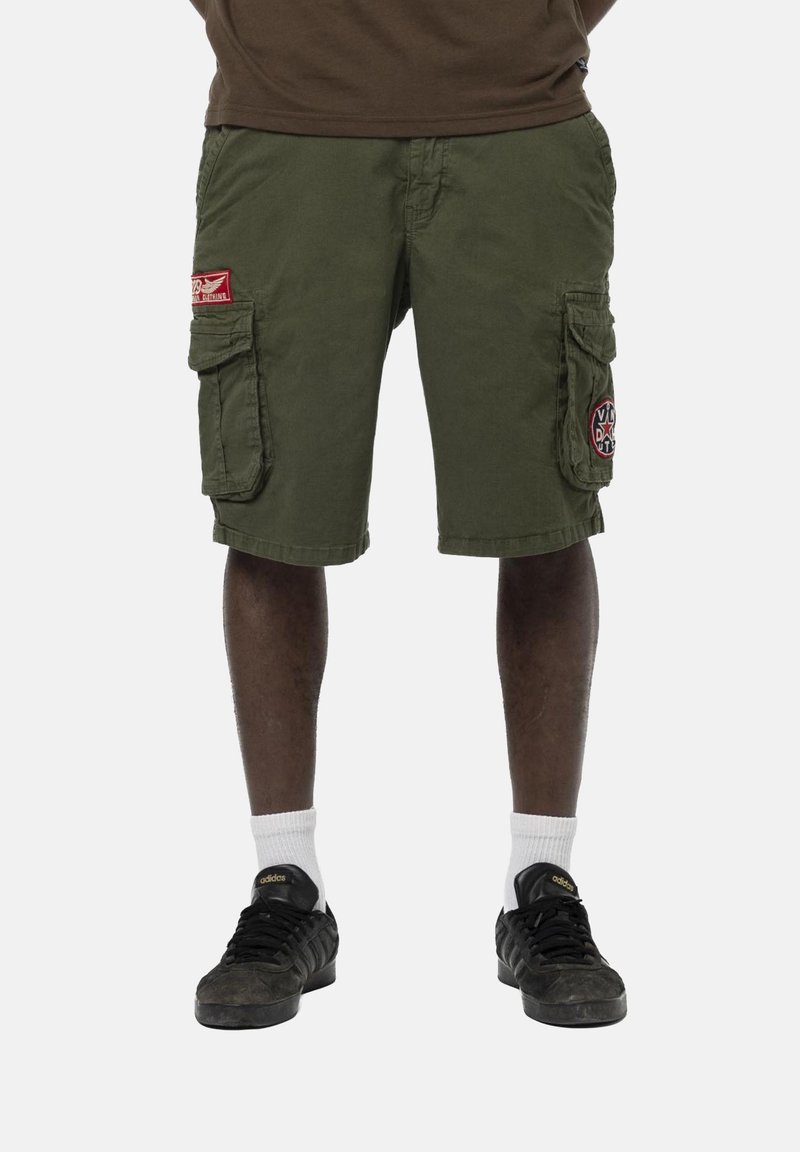 Homme portant un short cargo vert olive avec poches latérales, chaussettes blanches et baskets Adidas noires, debout contre un fond uni.