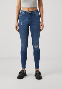 Jeans ajustados de mezclilla azul oscuro con un desgastado en una rodilla. Combinados con mocasines negros de plataforma con tachuelas plateadas.