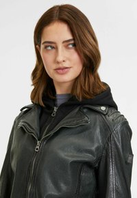 Zwarte leren motorjack met ritsen, metalen accenten en schouderdetails. Draagt boven een zwarte hoodie, met een gladde, gestructureerde afwerking.