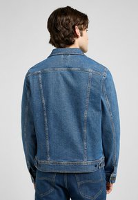 Chaqueta de mezclilla en azul medio. Presenta un cuello clásico, cierres de botón y detalles de costura. La textura es suave con un ligero acento de costura.