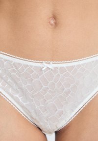 Hvite blonde truser med et geometrisk mønster, med bølgede kanter og en liten sløyfe detalj ved livet. Glatt, gjennomsiktig tekstur.