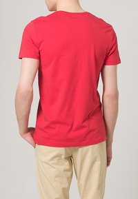 Rotes Baumwoll-T-Shirt mit Rundhalsausschnitt, kurzen Ärmeln und glatter Textur. Getragen in hellbeige Hosen.